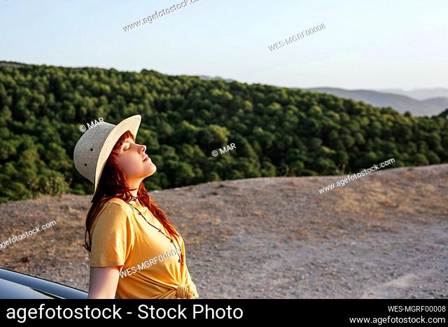 Relaxed young redhead woman leaning on car during road trip