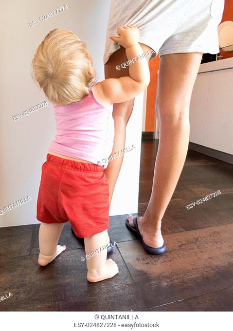 blonde baby red shorts clutching mom leg in white kitchen