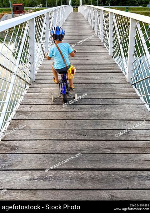 A boy with bicycle on a bridge. The boy has bicycle helmet. White suspension bridge over the river Drava