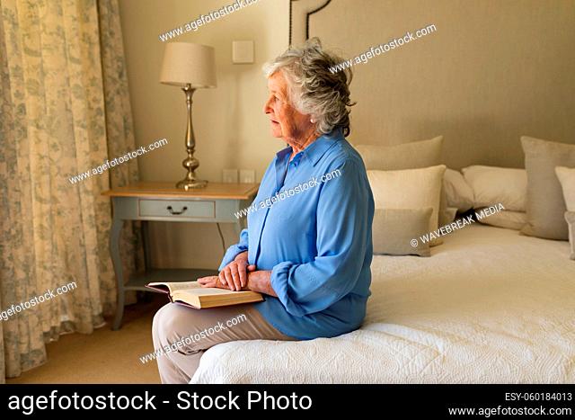 Senior caucasian woman sitting on bed and reading book in bedroom