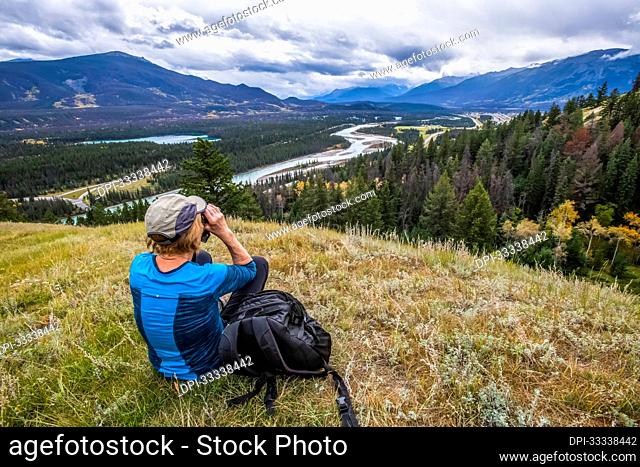 Woman on grassy knoll scans with binoculars up the Athabasca River Valley toward the town of Jasper in Jasper National Park; Alberta, Canada