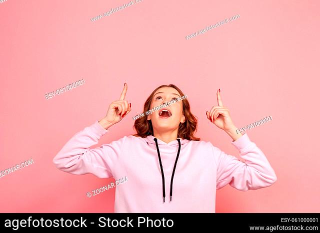 Portrait of surprised teenage girl in hoodie with open mouth pointing fingers up at empty copy space, place for advertisement