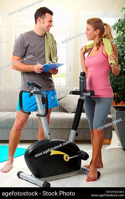 Young woman talking with her personal trainer standing beside exercise bike at home