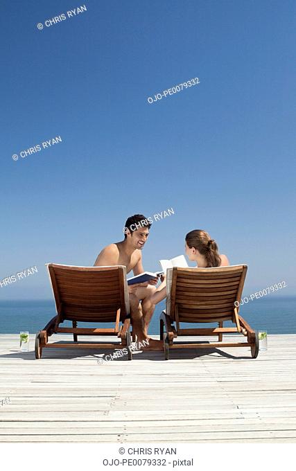 Couple in lounge chairs reading books near ocean