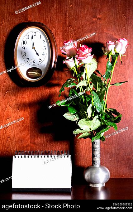 wall clock, blank calendar and pink roses in pewter jug on dark brown wooden background