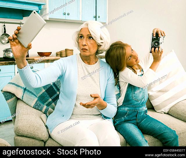 Grandma With Her Grandchild Use Each Other's Gadgets For Taking Selfie Pictures Senior Lady And Cheerful Girl Trying To Make Photo Session With Unusual Gadgets...