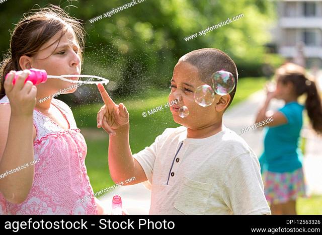 A girl stands blowing bubbles with a boy touching the bubble wand