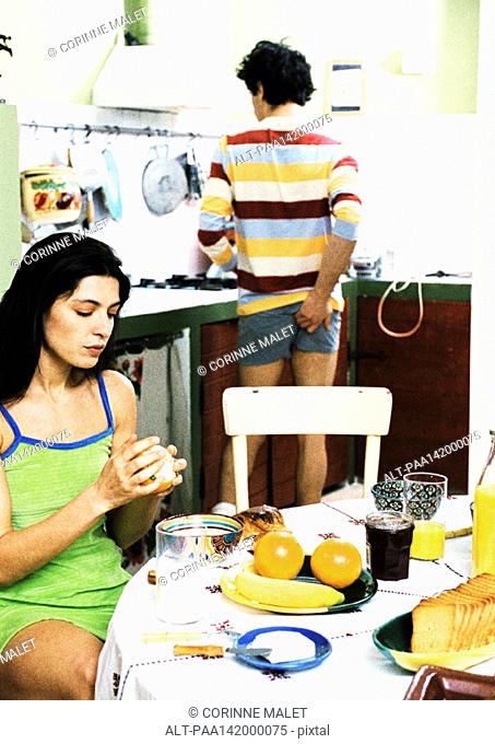Woman sitting at breakfast table, man standing, scratching rear
