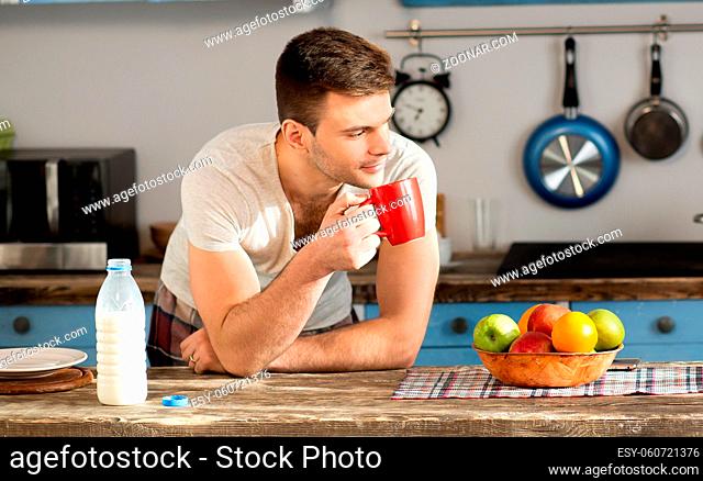 Close up view on a man with red cup in the kitchen. Young male standing, thinking and holding mug in his hand at home