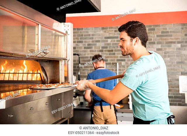 Side view of handsome young chef using peel to put pizza in oven for baking