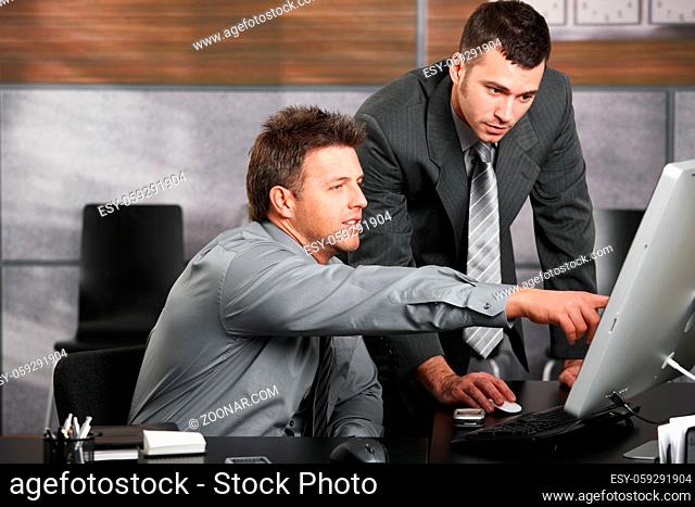 Two businessmen working together with computer at office desk, one of them pointing at screen