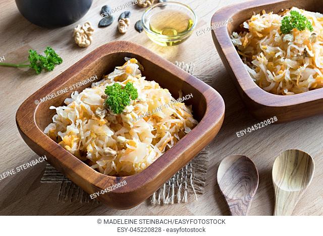 Fermented cabbage and carrots in two wooden bowls on a wooden table, with walnuts, parsley and pumpkin seeds in the background