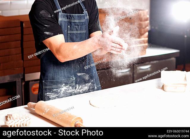 Roll out the dough. expanding cloud of flour. Closeup hand of chef baker in uniform blue apron cook pizza at kitchen