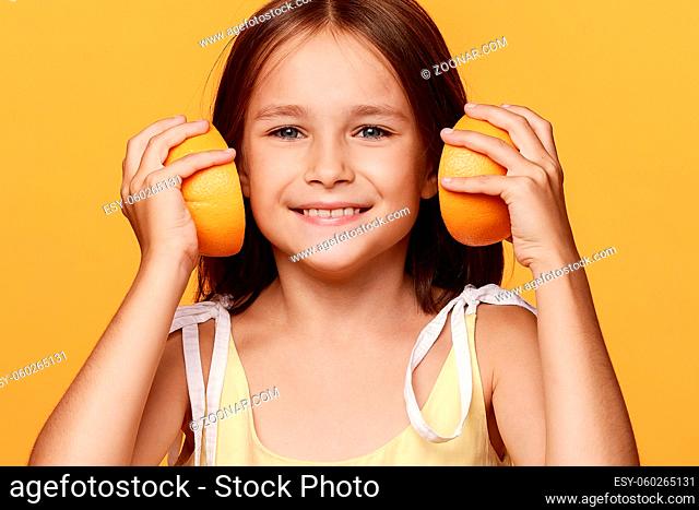 A little girl in a yellow dress, on an orange background, makes headphones out of orange halves. In the studio. Happy childhood. Fashion. Creative