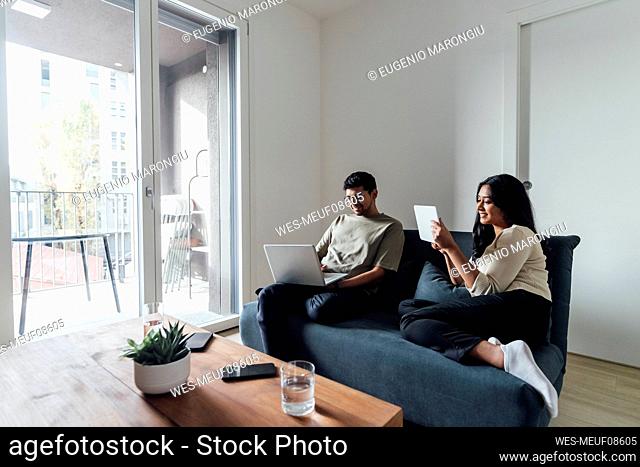 Young man and woman studying using laptop and tablet PC on sofa in living room
