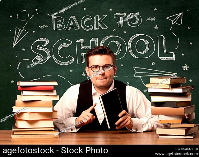 A young teacher in glasses sitting at classroom desk with pile of books in front of blackboard saying back to school drawing concept