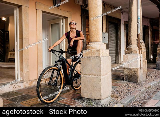 Smiling woman looking away while standing on mountain bike by pillar on street