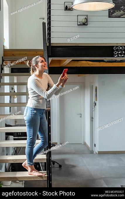 Smiling woman controlling home functions with smart phone while standing on steps at home