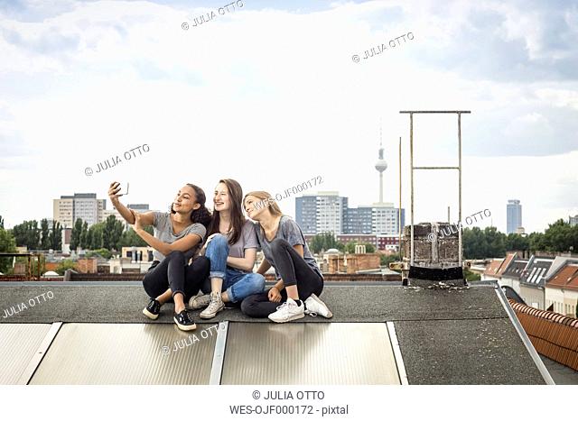 Germany, Berlin, three friends sitting side by side on roof top taking selfie with smartphone
