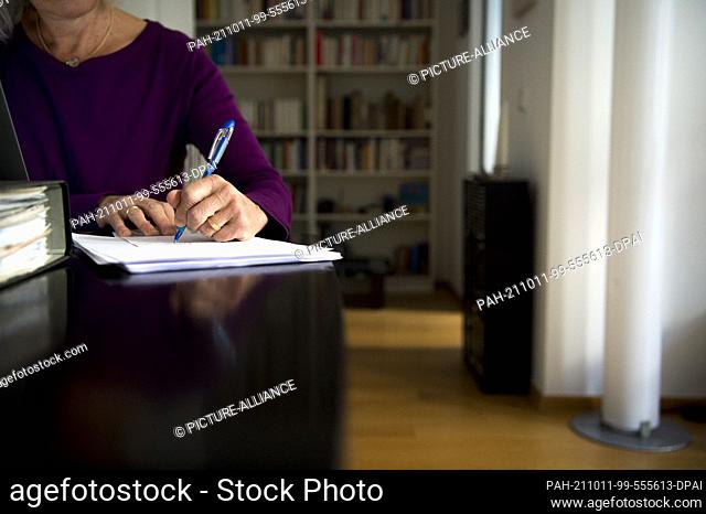 PRODUCTION - 01 October 2021, Bavaria, Munich: A woman is sitting at a dining table at home. She is writing with a pen on a piece of paper next to a laptop