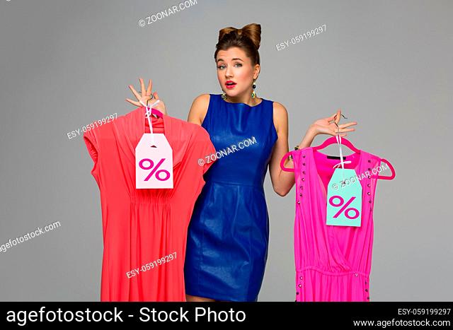 Beautiful confused woman holding two dresses on hangers. Price tags. Over grey background. Copy space