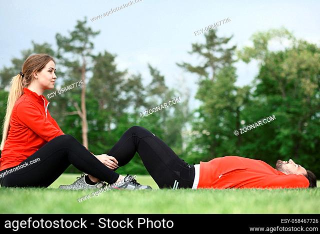 Sport man and woman training in green park or forest all together. Lady holding man#39;s legs who is doing abdominal crunches