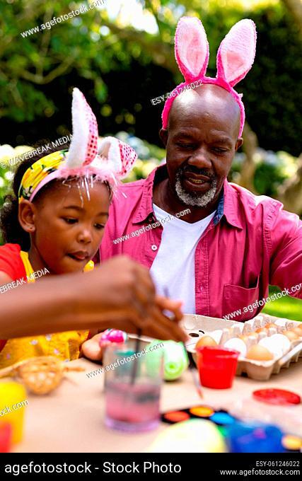 African american girl and grandfather in bunny ears painting eggs on easter day