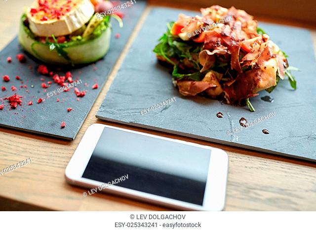 food, eating and technology concept - goat cheese and prosciutto ham salads on stone plates with smartphone at restaurant or cafe