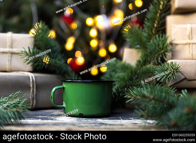 Coffee in a green tin mug on the rustic table with wrapped gifts, Christmas tree lights and spruce branches in the background