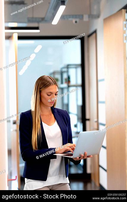 Smartly dressed caucasian businesswoman standing in corridor using laptop computer