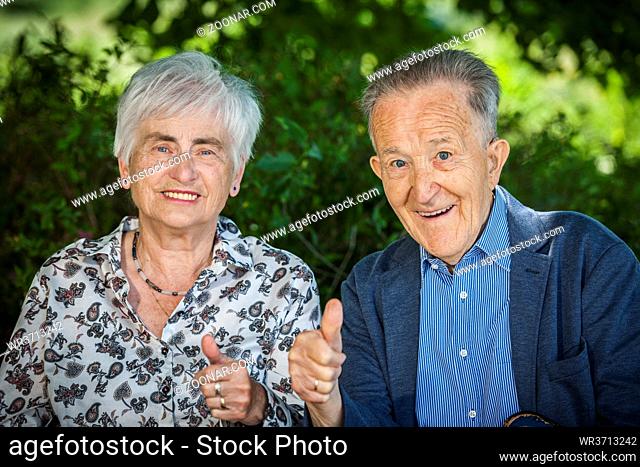 Head-and-shoulder portrait of an 80 year old retired couple in front of green plants background showing the thumbs up and friendly looking at the camera