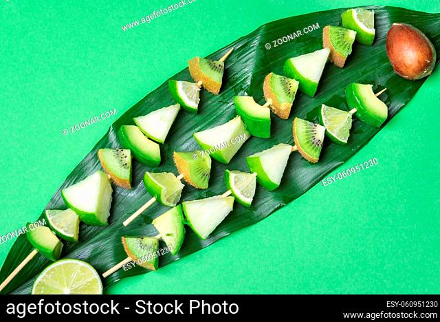 Above view of delicious green fruits skewers on a big leaf, on a green tabletop. Minimalist vegetarian food. Detox meal. Healthy diet desserts