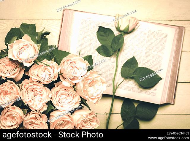 Vintage image with a bouquet of roses in the foreground and an old open book with a single rose on it in the background, placed on a wooden table