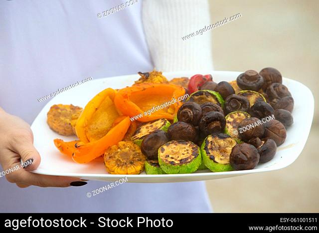 Hands hold a plate of grilled vegetables and mushrooms