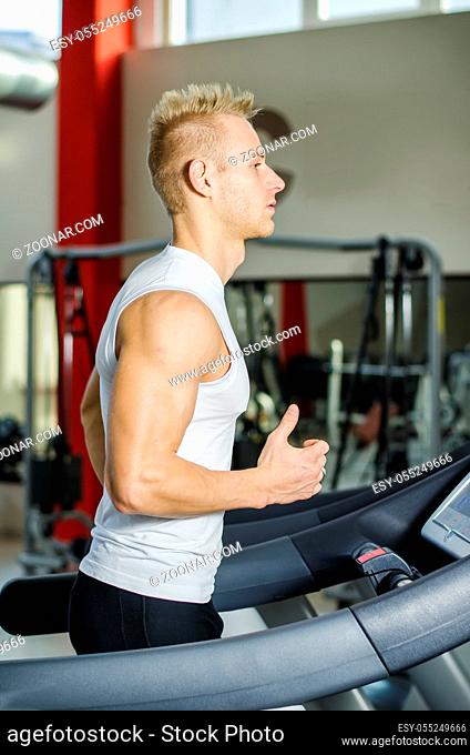 Blond attractive young man running on treadmill, seen from a side. Profile shot of athlete