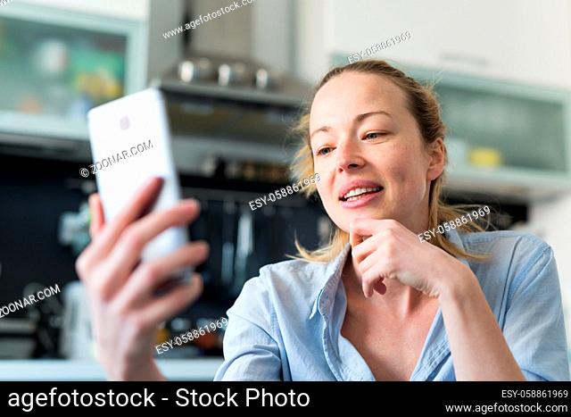 Young smiling cheerful woman indoors at home kitchen using social media apps on phone for video chatting and stying connected with her loved ones