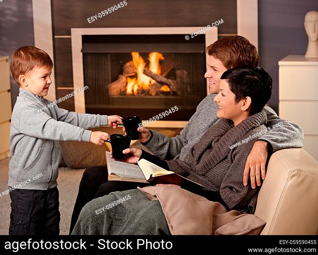 Happy family sitting on couch at home in front of fireplace, drinking tea, smiling
