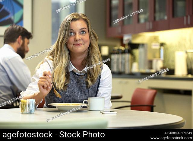Young Caucasian woman looking at camera with a smile and exaggerated big eyes, while sitting in break area of office with lunch of soup and Hot Tea