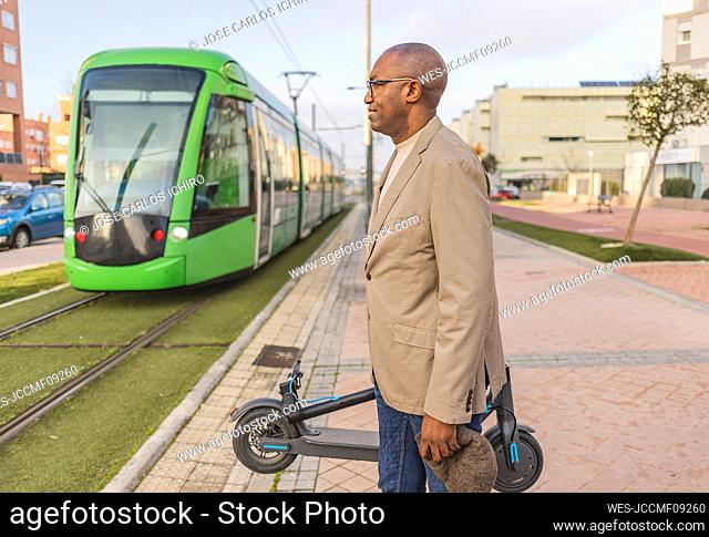 Businessman with electric push scooter waiting for tram on footpath