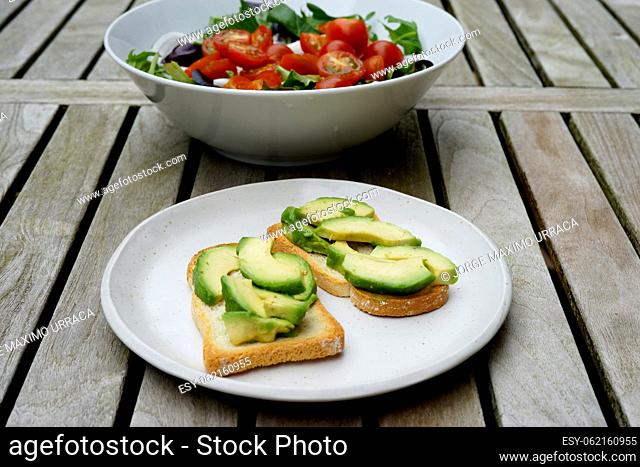 Toast with avocado on a white plate and a bowl of lettuce with cherry tomato