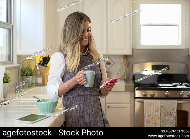 Young woman in her kitchen eating soup and drinking tea, smiling and laughing while checking smartphone