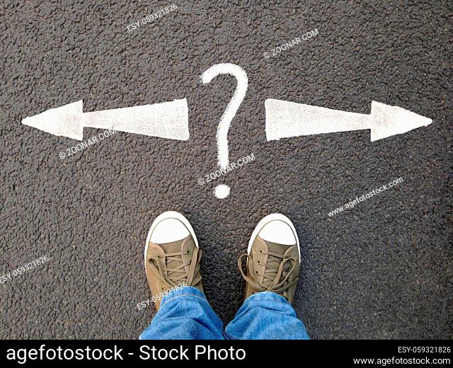 feet in canvas shoes standing on asphalt from personal perspective, road markings with arrows pointing left and right with question mark