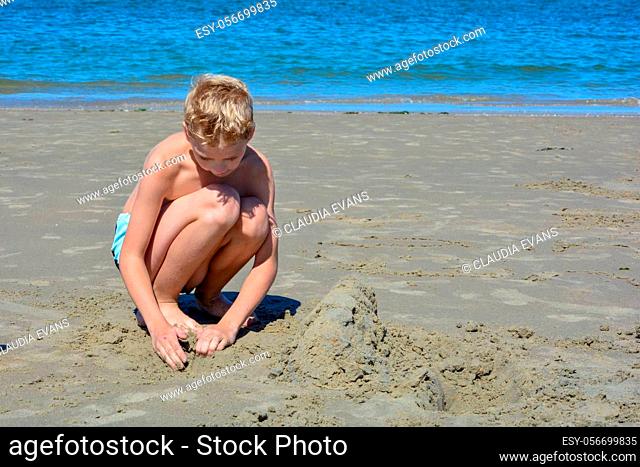 Little blond boy with swimming trunks builds a sand castle in the sand on the beach in front of the sea