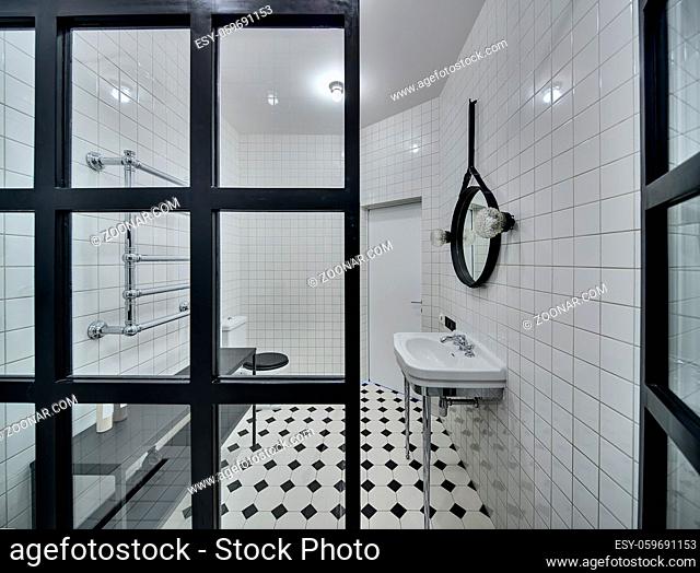 Modern bathroom with walls of white tiles. There is white washbasin, black mirror, lamps, glass partition, white-black toilet, black rack with accessories