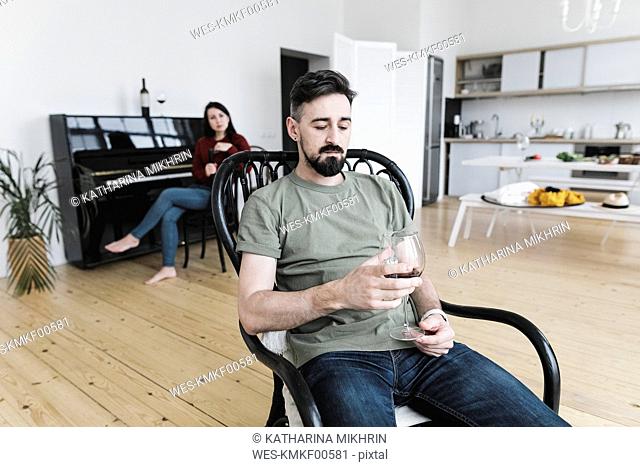 Couple at home, Man drinking wine, woman playing he piano in background