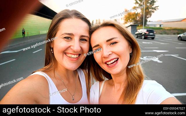 Closeup photo of brunette and blonde smiling girls making selfie on smartphne at car parking