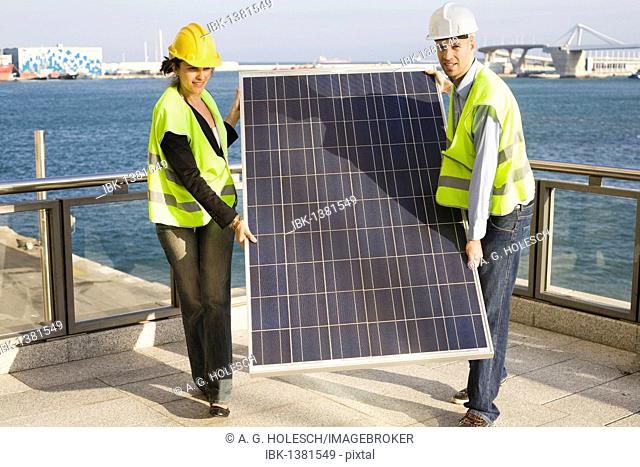 Two people in hard hats and protective jackets carrying a giant solar panel