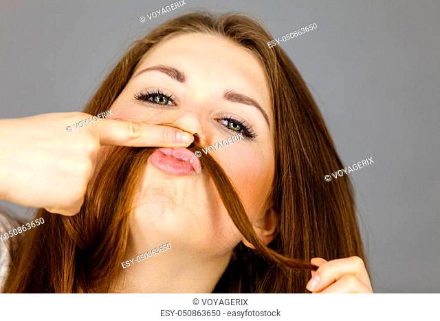 Woman having fun with her long brown hair making moustache being happy about hairdo condition, studio shot grey background