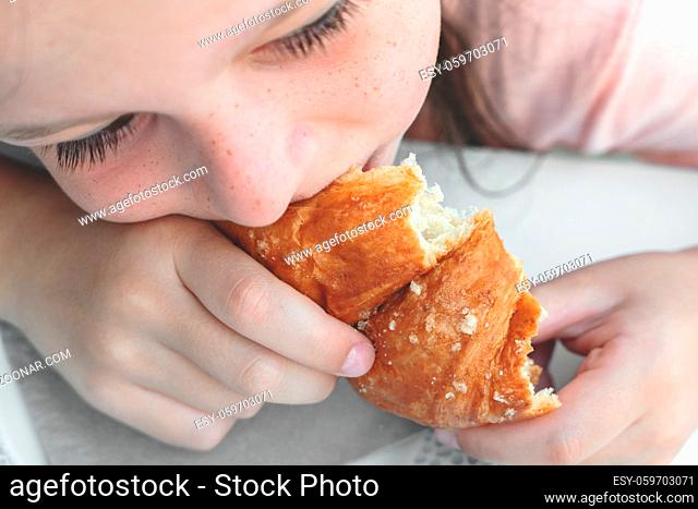 Pretty little girl eats a fresh croissant for breakfast. Portrait of cute girl with yummy face enjoy eating croissant. Healthy food for children concept