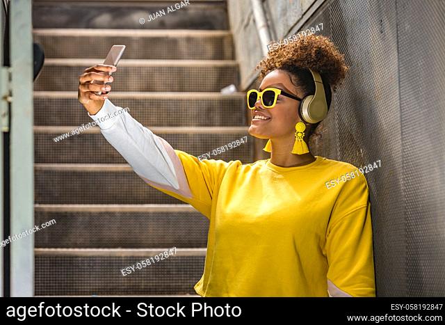 Happy millennial African American female in vivid yellow wear and trendy sunglasses taking selfie on mobile phone while listening to music with headphones on...